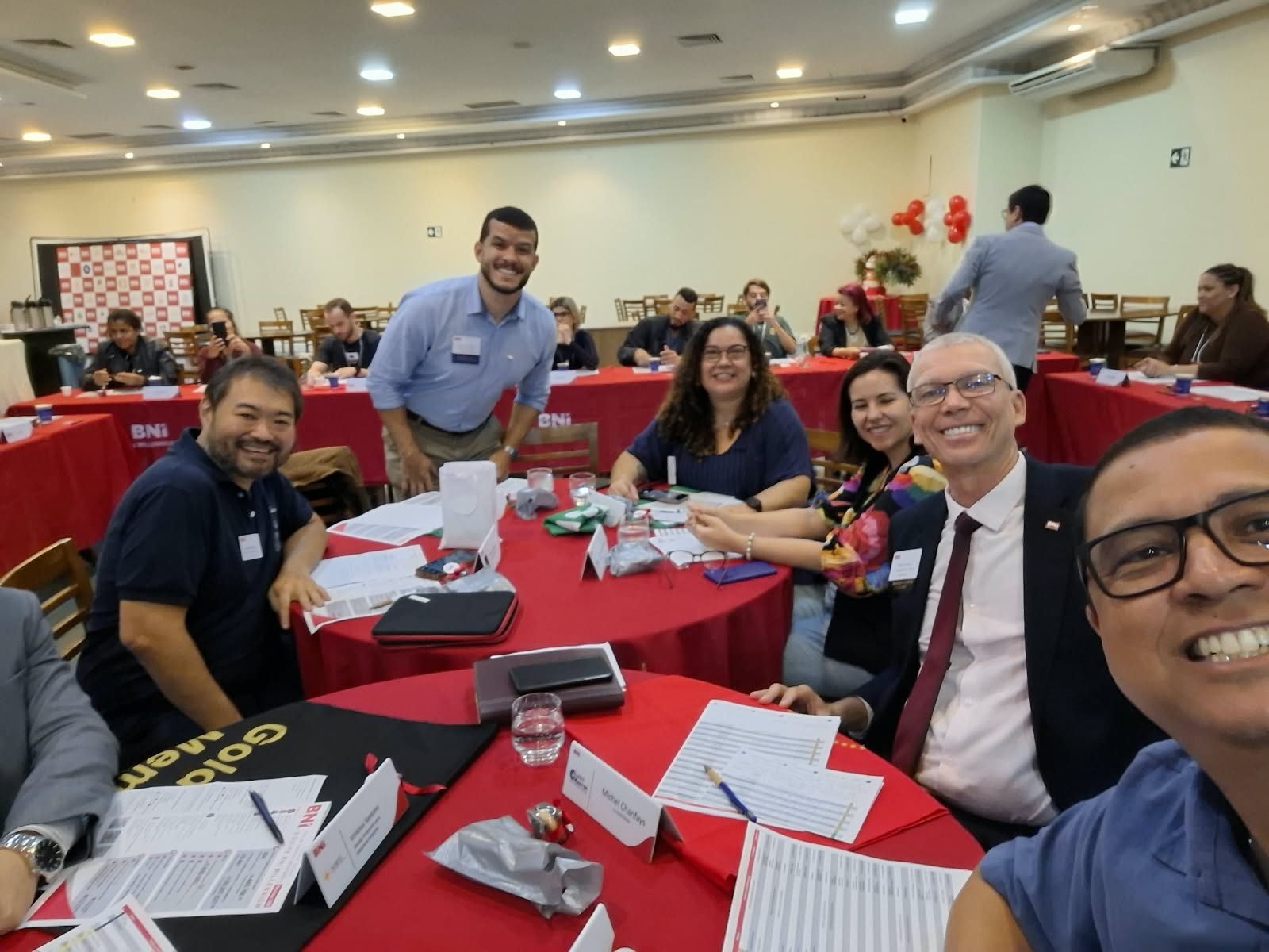 Participantes em volta de uma mesa redonda, com papéis sobre a toalha vermelha.