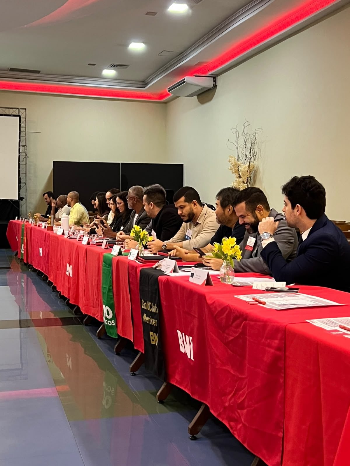 Vista da sala de reunião, com participantes sentados ao longo de uma mesa longa coberta de vermelho.